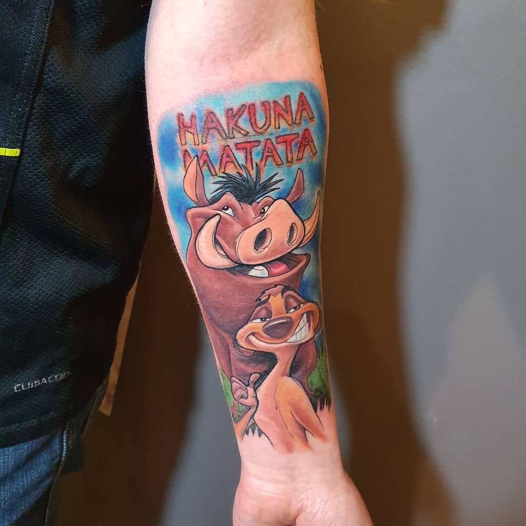 Forearm tattoo of Pumbaa and Timon from The Lion King with "Hakuna Matata" text above them